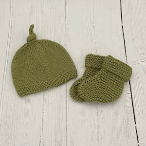 May include: A set of knitted baby clothes in a muted green color. The set includes a hat with a knotted top and a pair of booties.