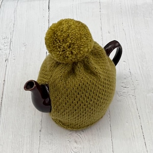 Op de afbeelding: Een bruine keramische theepot met een donkerbruin handvat en tuit is bedekt met een gebreide olijfgroene theemuts. De theemuts heeft een grote olijfgroene pompon bovenop. De theepot staat op een wit houten oppervlak.
