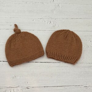 Peut inclure: Deux bonnets en tricot marron, l'un avec un sommet noué, sur un fond en bois blanc.