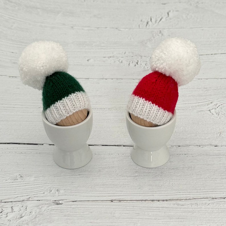 May include: Two knitted egg cozies in egg cups. One is red with a white pom pom and the other is green with a white pom pom.