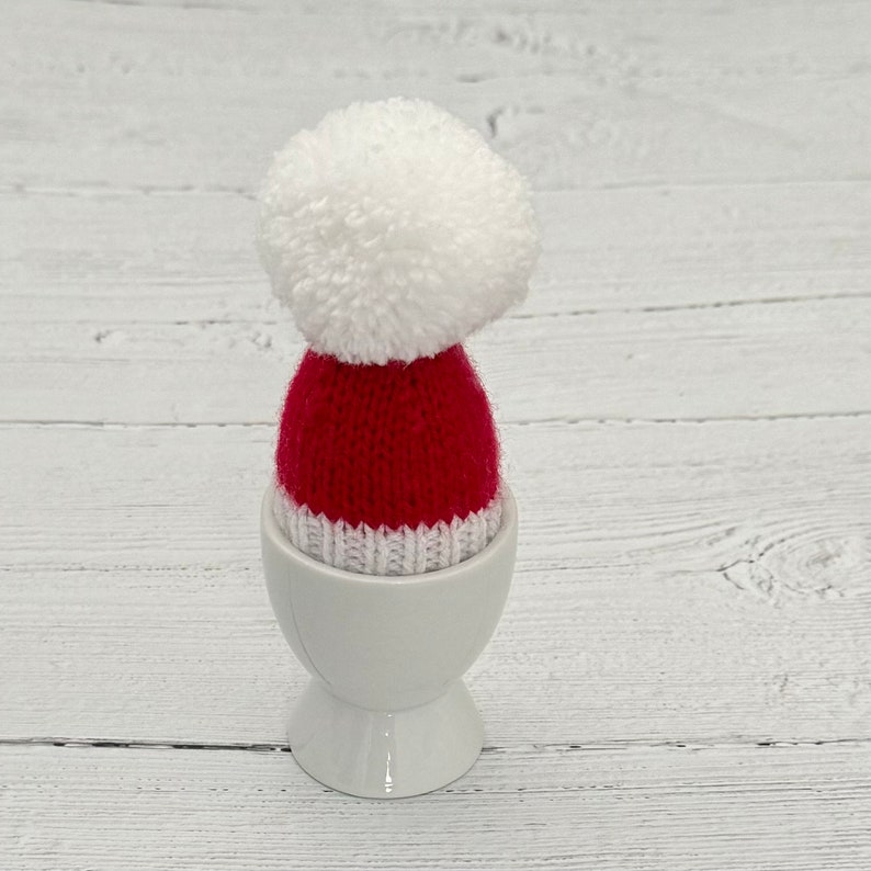 May include: A white ceramic egg cup with a red and white knitted Santa hat with a white pom-pom on top.
