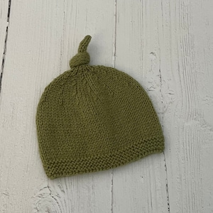 May include: A green knitted hat with a knotted top. The hat is made of a soft, textured yarn.