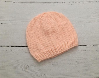 May include: A peach-coloured knitted beanie hat. The hat is made of a soft, textured yarn and has a simple, classic design.