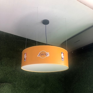 May include: A yellow round ceiling light fixture with the Los Angeles Lakers logo and NBA logo. The light fixture is hanging from the ceiling by three wires.