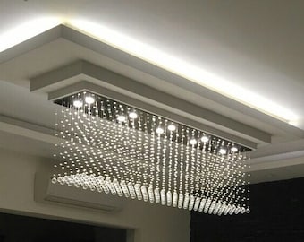 May include: A modern chandelier with a rectangular frame and cascading crystal strands. The chandelier is suspended from a white ceiling with recessed lighting.
