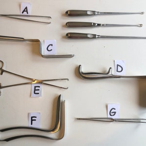 Puede incluir: Un conjunto de siete instrumentos quirúrgicos, incluyendo fórceps, un espéculo y un gancho, dispuestos sobre una superficie blanca. Los instrumentos están etiquetados con las letras A a G.