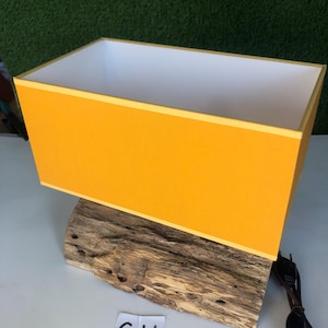 Puede incluir: Una pantalla de lámpara rectangular amarilla con un interior blanco se asienta sobre un trozo de madera a la deriva. La lámpara tiene un cable y un enchufe negros. La madera a la deriva tiene las letras "GH" escritas en ella.