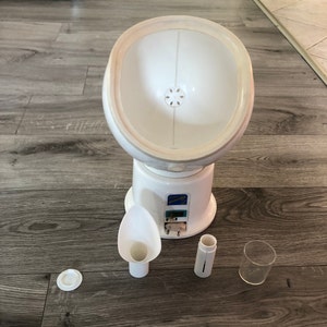 May include: White facial steamer with a round, white bowl, a white spout, and a white tube. The steamer is on a wooden surface. The steamer has a blue label that says "Home Professional".