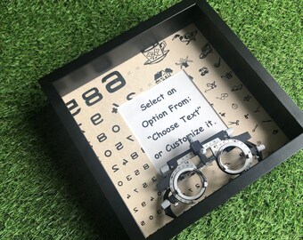 May include: A black shadow box with a brown background featuring an eye chart, a vintage eye exam tool, and a white card with the text "Select an Option From: "Choose Text" or Customize it."