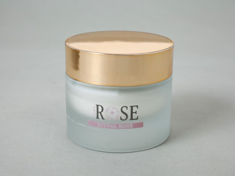 May include: A cylindrical glass jar with a frosted finish and a shiny gold lid. The front of the jar features the text "EDEN ROSE" in black lettering with a rose graphic replacing the letter "O", and the text "RITUAL ROSE" on a pink bar below.