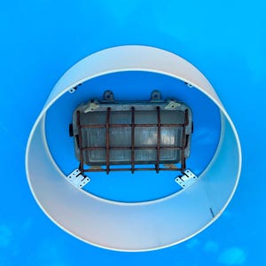 Op de afbeelding: Een vintage industriële lamp met een rechthoekige lamp in een kooi. De lamp is omhuld door een roestige metalen kooi en gemonteerd op een blauwe achtergrond. Een witte, cirkelvormige kap omringt de lamp, wat een uniek ontwerp creëert.