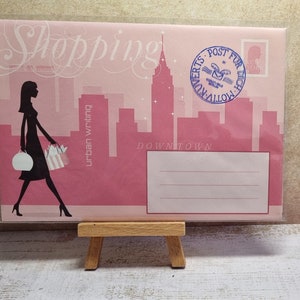 May include: Pink and white shopping bag with the text "Shopping urban writing Downtown" and a silhouette of a woman carrying shopping bags. The bag has a blue stamp with the text "Post fur Dich" and a small image of a building.