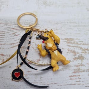 Op de afbeelding: Een goudkleurige sleutelhanger met een gele hondenfiguur met een kroon en een zwart vest. De sleutelhanger bevat zwarte en gouden linten, een hartvormige bedel en een kleine kroonbedel.