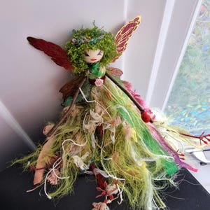 May include: A handmade fairy doll with a wooden head, green yarn hair, and red and gold wings. The fairy is dressed in a green and yellow dress with floral accents and a ladybird detail. A whimsical decorative item.