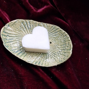 Può includere: Un portasapone in ceramica fatto a mano con un sapone a forma di cuore. Il piatto ovale ha una superficie strutturata verde chiaro e un piccolo foro di drenaggio. Il sapone bianco è al centro.