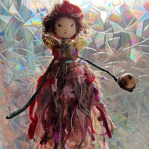 May include: A handcrafted fairy doll with a painted wooden face, a red hat, and a fabric dress in shades of red, pink, and brown. The doll has gold wings and holds a small, round object on a stick.