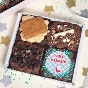 May include: A white cardboard box containing four square chocolate brownies with various toppings. One brownie features a round edible topper with a blue checkered pattern, the text YOU PASSED!, and a red L symbol. Others include a biscuit, chocolate pieces, and cookie crumbles.
