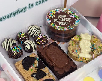 May include: A white box filled with a selection of chocolate treats, including a small round cake with sprinkles and a candle, a brownie with chocolate frosting, a cookie with white chocolate sprinkles, and chocolate-covered truffles. The box is decorated with the words "Ruby, The Cake Artist" and "everyday is cake day..."