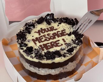 Puede incluir: Un pastel redondo de chocolate con glaseado blanco y migas de galletas Oreo. El glaseado tiene las palabras "YOUR MESSAGE HERE!" escritas en chocolate. El pastel está en una caja blanca con una tapa transparente.