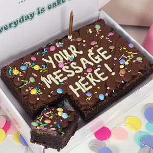 May include: A large chocolate brownie with a personalised message written in white chocolate. The brownie is decorated with colourful sprinkles and chocolate sweets. The brownie is in a white box with a pink and green design. The text on the box reads "The everyday is cake".