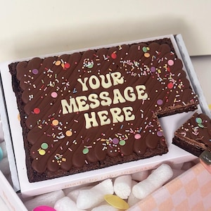 May include: A large chocolate brownie with the words "YOUR MESSAGE HERE" in white icing. The brownie is decorated with chocolate lines and colorful sprinkles. It is presented in a white box with pink and white checkered paper.