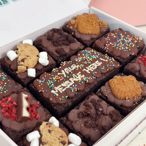 May include: A white box filled with nine different chocolate brownies. Each brownie is topped with a variety of toppings, including sprinkles, chocolate chips, cookies, and marshmallows. The box is labeled with the text "Your Message Here".