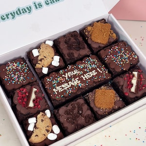 May include: A white box filled with nine chocolate brownies, each topped with different sweet treats like cookies, marshmallows, sprinkles, and chocolate. The box has the text "everyday is cake" on the top and one brownie has the text "Your message here!" written in white icing.