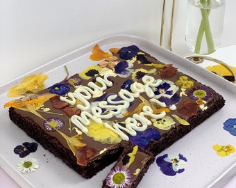 Puede incluir: Un brownie cuadrado de chocolate cubierto con flores comestibles y escritura de chocolate blanco que dice "your message here!" El brownie está en un plato blanco con más flores esparcidas a su alrededor.