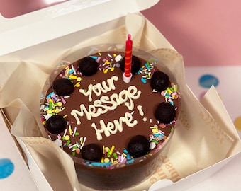 May include: A small chocolate cake with a single red and white striped candle in a white box. The cake is decorated with colorful sprinkles and chocolate balls. The cake has the words "Your Message Here!" written in white icing.