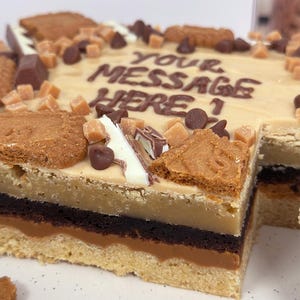 K&ouml;nnte beinhalten: Ein mehrschichtiger Kuchen mit einer braunen Zuckerkeks-Kruste, einer Schicht Schokoladen-Brownie, einer Schicht Karamell und einer Schicht braunen Zuckerkeksteig. Der Kuchen ist mit einer Schicht braunen Zuckerkekskr&uuml;meln, Schokoladenst&uuml;ckchen, Karamellst&uuml;cken und einer wei&szlig;en Schokoladentafel bedeckt. Der Kuchen ist mit den Worten "Ihre Nachricht hier!" in braunem Zuckerguss verziert.