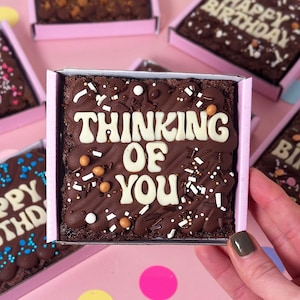 May include: A chocolate brownie in a pink box, decorated with white icing spelling "THINKING OF YOU". The brownie is covered in sprinkles and small candies. Other brownies with text, such as "HAPPY BIRTHDAY", are visible in the background.