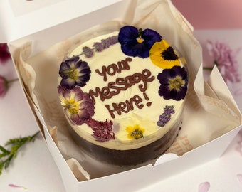 Puede incluir: Un pastel blanco con glaseado de chocolate y flores comestibles. El glaseado tiene las palabras "Your Message Here!" escritas en él.