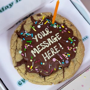 May include: A large chocolate chip cookie topped with melted chocolate and colorful sprinkles. The chocolate has the words "YOUR MESSAGE HERE!" written in white icing.