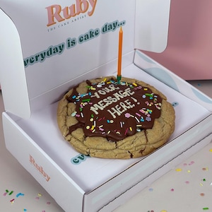 May include: A large chocolate chip cookie with chocolate icing and colorful sprinkles. The cookie has a personalized message written in chocolate icing that says "YOUR MESSAGE HERE!" A single orange candle is stuck in the cookie. The cookie is in a white box with the words "Ruby THE CAKE ARTIST" and "everyday is cake day..." printed on the box.