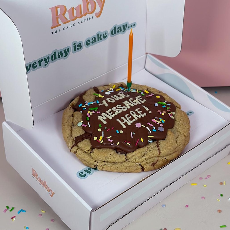 May include: A large chocolate chip cookie with chocolate icing and colorful sprinkles. The cookie has a personalized message written in chocolate icing that says "YOUR MESSAGE HERE!" A single orange candle is stuck in the cookie. The cookie is in a white box with the words "Ruby THE CAKE ARTIST" and "everyday is cake day..." printed on the box.