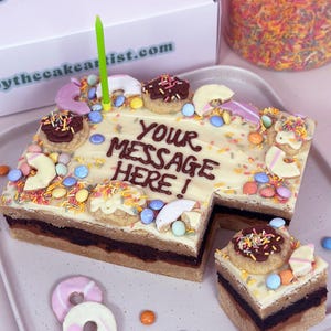 May include: A multi-layered cake with a white chocolate frosting and colourful sprinkles. The cake is shaped like a house and has a message written in chocolate icing that says "YOUR MESSAGE HERE!"