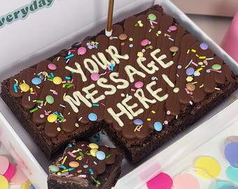 Op de afbeelding: Een rechthoekige chocoladebrownie in een witte doos, bedekt met chocoladeglazuur, kleurrijke hagelslag en chocoladesnoepjes. De tekst "YOUR MESSAGE HERE!" is geschreven in wit glazuur en er staat een gouden kaarsje op de brownie.