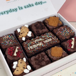 May include: A white box with a lid open, revealing a variety of chocolate brownies with different toppings. The brownies are arranged in a grid pattern and include toppings like sprinkles, cookies, and chocolate chips. The box has the text "Everyday is cake day..." printed on the lid.