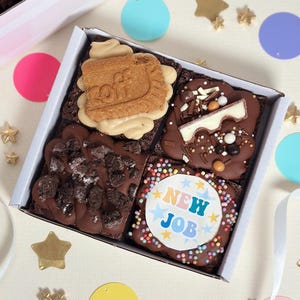 May include: A square white box containing four chocolate brownies with distinct toppings: one with a tan biscuit, one with chocolate shavings and a white chocolate piece, one with dark biscuit crumbles, and one with a round edible topper reading NEW JOB.