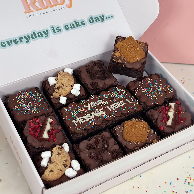May include: A white box with the text "The Cake Artist" and "everyday is cake day..." on the lid. The box is open and contains nine different brownie squares with various toppings, including sprinkles, chocolate chips, cookies, and marshmallows.
