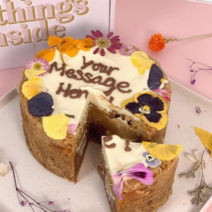 May include: A round cake with a white frosting base and a chocolate message that says "Your Message Here". The cake is decorated with edible flowers and a slice has been cut out.