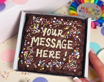 Puede incluir: Un brownie de chocolate cuadrado en una caja blanca, cubierto con remolinos de glaseado de chocolate negro, virutas de colores y texto en glaseado blanco que dice "YOUR MESSAGE HERE !". El brownie tiene un borde de glaseado de chocolate.