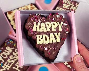 May include: A heart-shaped chocolate brownie with pink and gold sprinkles and white icing that says "Happy Bday".