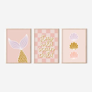 以下が含まれることがあります： 人魚の尾、貝殻、そして「Stay Wild Ocean Child」のテキストが描かれた、額装された3つのアートプリントのセット。プリントはピンク、ゴールド、ラベンダーの色使いです。人魚の尾は金色の鱗模様です。