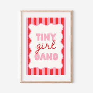 Può includere: Una stampa artistica incorniciata con le parole "TINY girl GANG" in rosa e rosso. Il testo è su uno sfondo bianco con un bordo ondulato, incorniciato da strisce verticali rosse e rosa. La cornice è color legno chiaro.