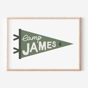 以下が含まれることがあります： 白い文字で「camp JAMES」と小さな松の木のグラフィックが書かれた緑色のペナント。ペナントは明るい木製のフレームで囲まれています
