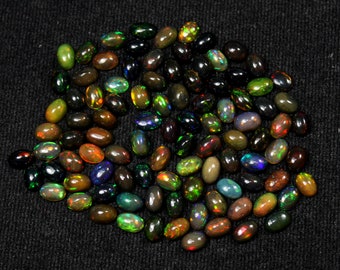 May include: A collection of oval-shaped black opal gemstones. The opals display a variety of colors, including green, blue, red, and yellow, with flashes of iridescence. The gemstones are scattered on a dark, textured surface.