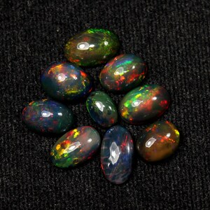 May include: A collection of nine oval-shaped black opals with vibrant internal color flashes of red, green, and yellow. The opals are arranged on a dark gray, textured surface, showcasing their iridescent qualities.