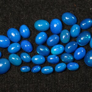 May include: A collection of oval-shaped, polished gemstones in varying shades of blue, from light to deep teal. The gemstones are scattered on a dark, textured surface, showcasing their glossy finish and internal iridescence.