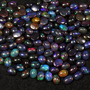 May include: A collection of polished black opal gemstones. The opals have a dark base color with flashes of iridescent colors, including green, blue, and orange. The gemstones are oval-shaped and vary in size, scattered on a dark surface.